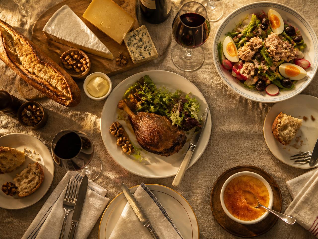 A French table set with baguette, cheeses, duck confit, salad, red wine and crème brûlée